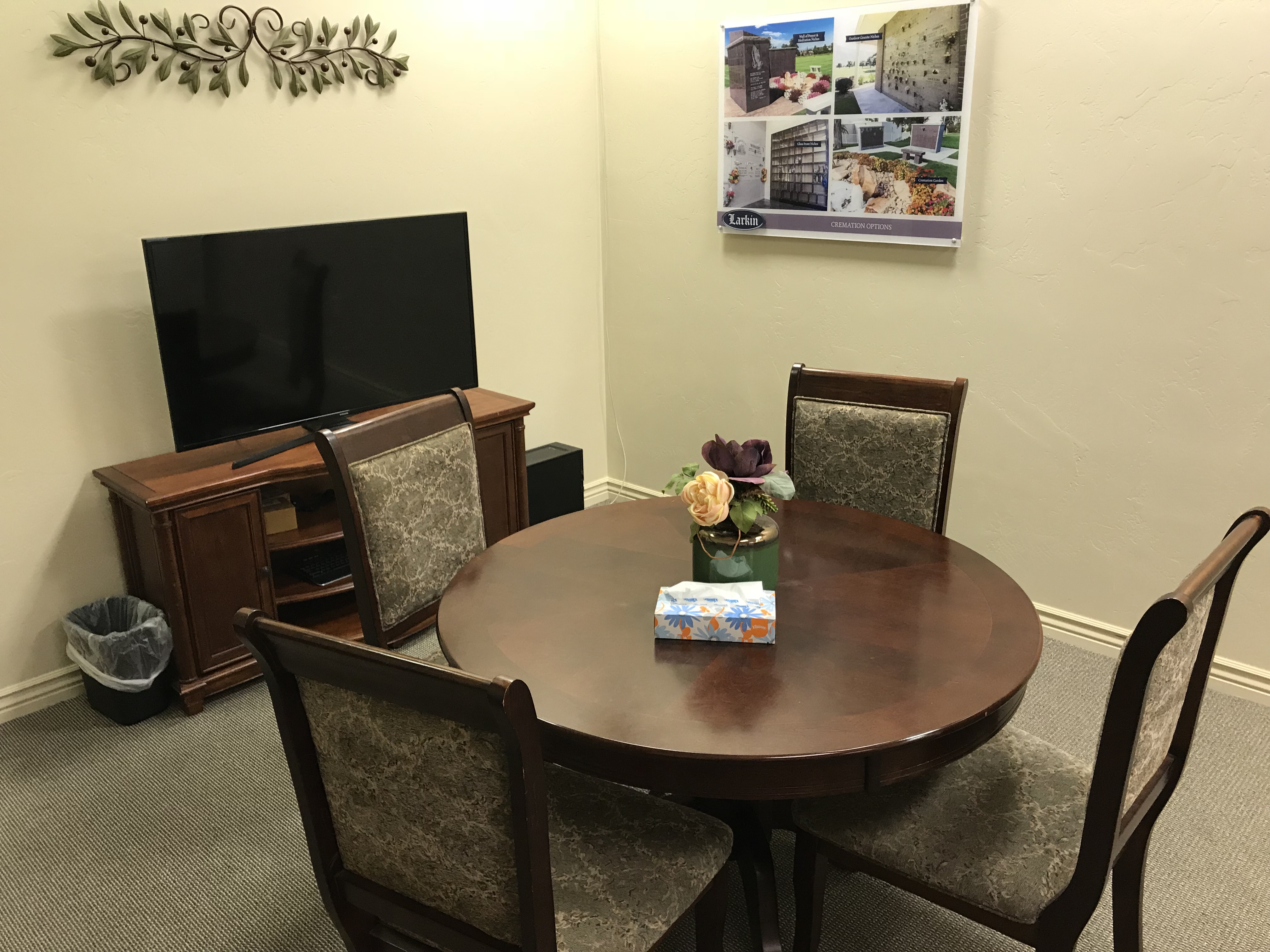 Arrangement room with a round dark-wood table, four upholstered chairs, a small floral arrangement and a wall screen for reviewing urn options.