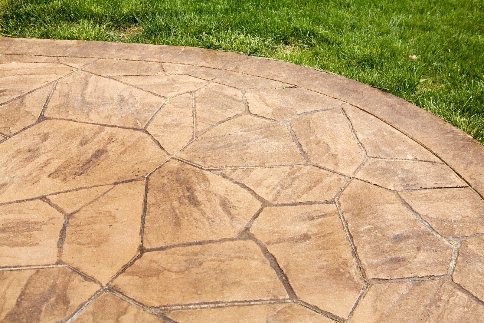 A close-up view of a textured, tan-colored stamped concrete patio or walkway with a flagstone pattern and a curved edge bordering green grass.