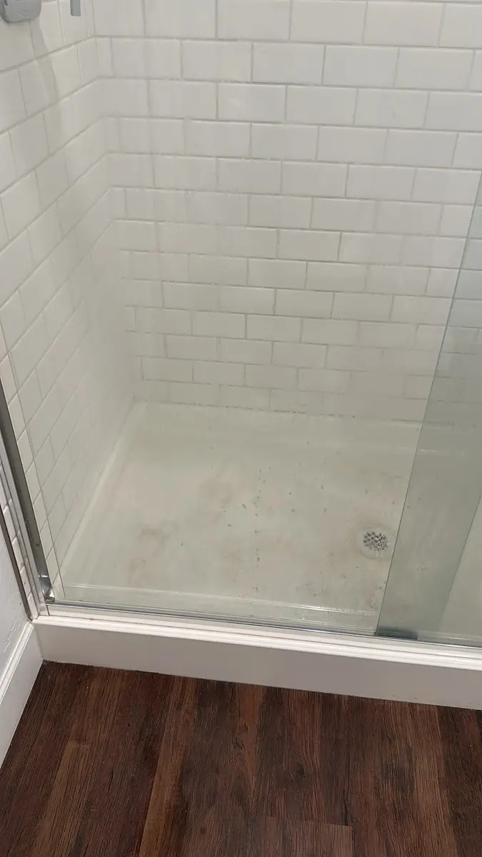 A close-up view of a shower stall with white subway tile on the walls and a light-colored shower pan. The shower pan appears to have some stains and debris. A clear glass shower door is partially visible, and dark wood-look flooring is visible in the foreground.