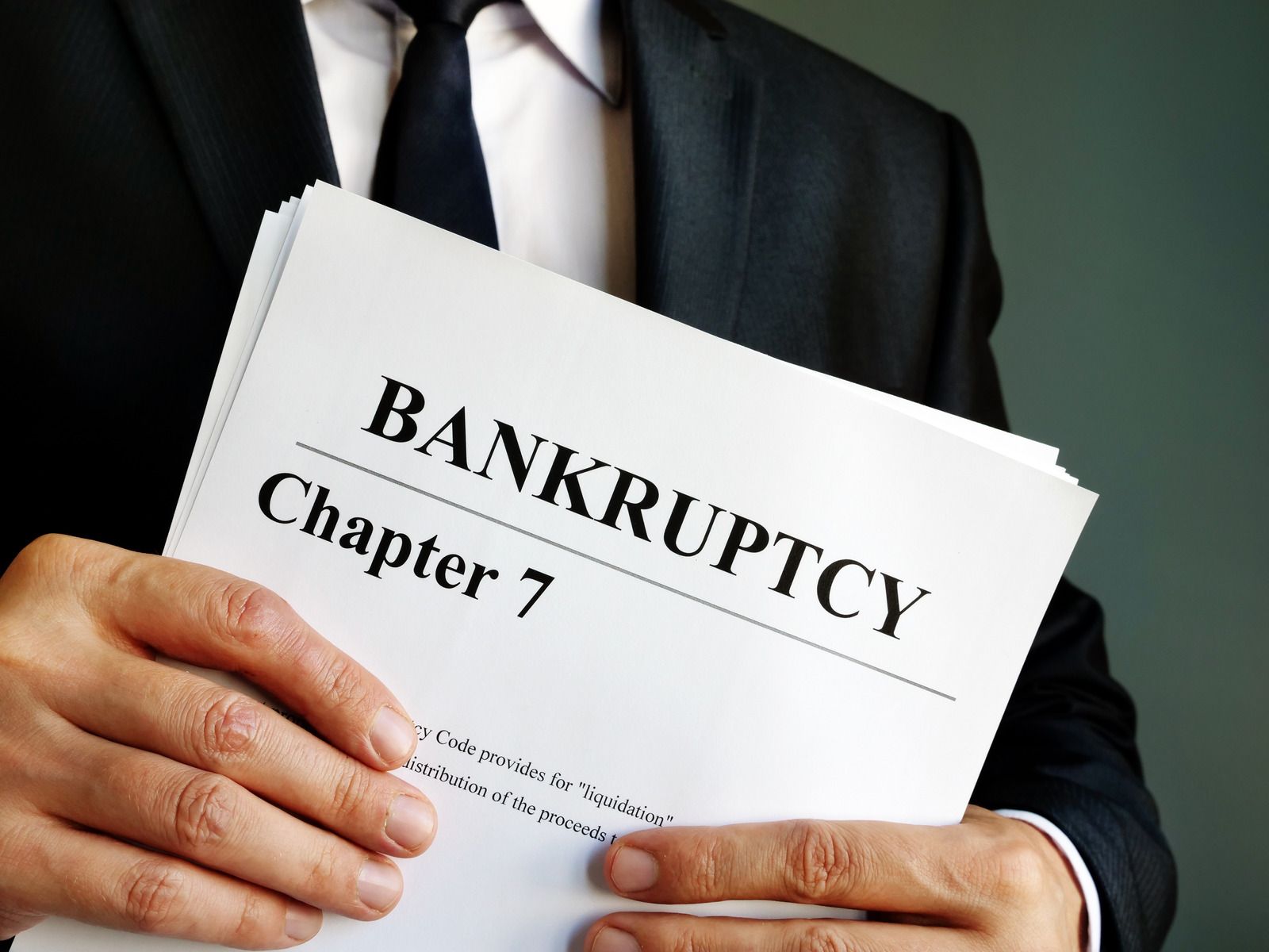 A person in a dark suit, white shirt, and black tie holds a stack of papers titled "Bankruptcy Chapter 7," with text about liquidation and distribution of proceeds visible.