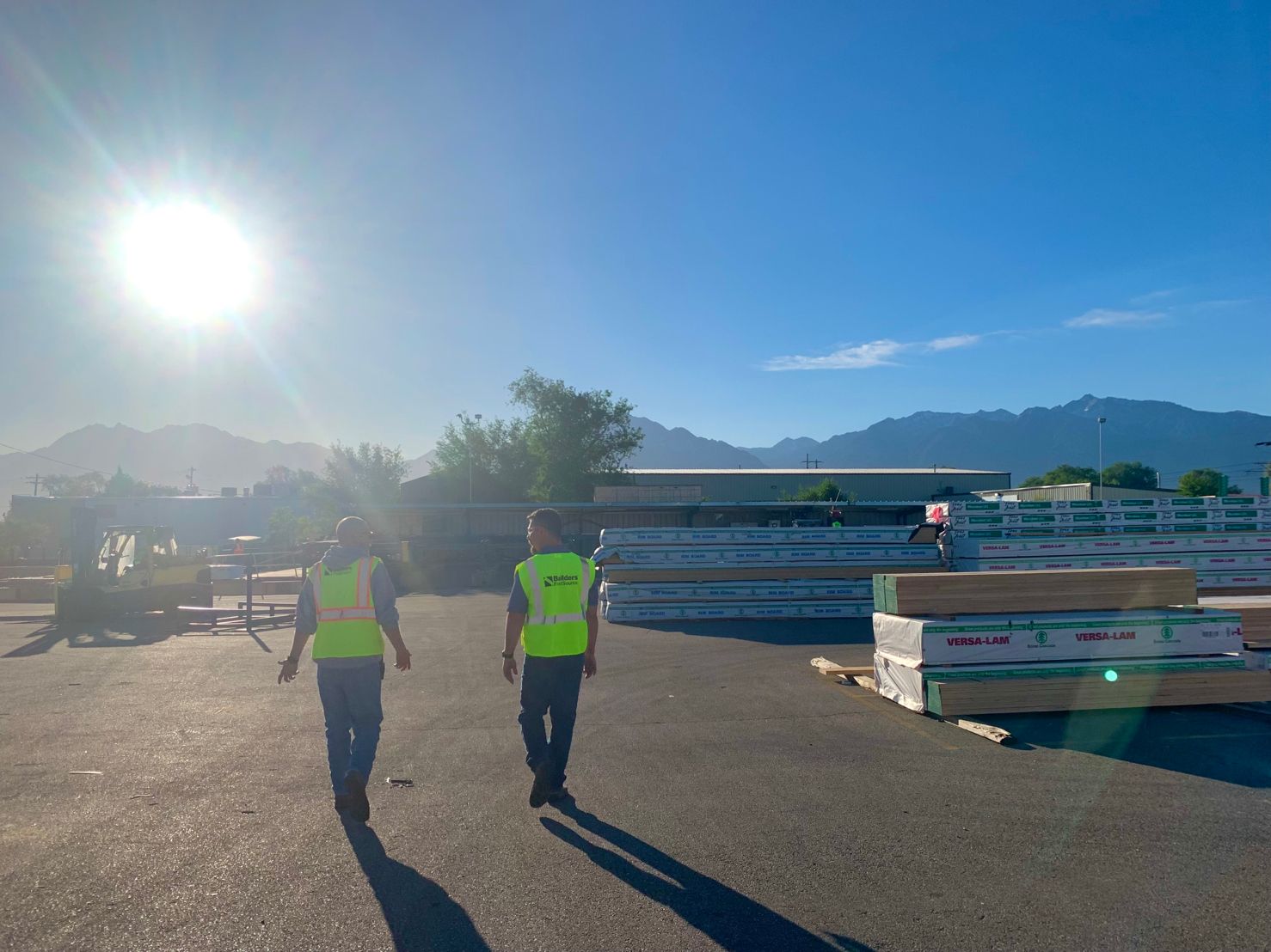 Two employees in safety vests walking through a lumber yard at the Builders FirstSource READY-FRAME facility. The yard is filled with stacks of Versa-Lam and other building materials, with mountain peaks visible in the background under a bright morning sun.
