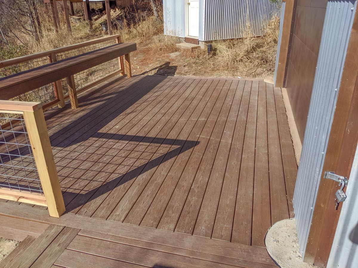 Embrace rustic charm with this custom composite deck by DeckPro LLC. Designed to blend perfectly with the natural surroundings and the building's corrugated siding, it features durable brown composite decking. The unique wood and industrial wire mesh railing offers safety without blocking the view, alongside a convenient built-in bench for relaxing outdoors.