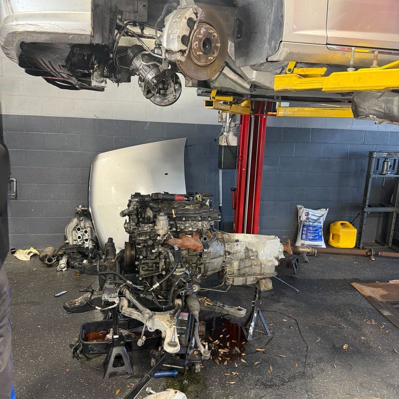 This image shows a vehicle lifted high on a yellow hydraulic lift in a garage. Below the vehicle, its engine, transmission, and various components are detached and resting on the concrete floor, indicating extensive automotive repair work.