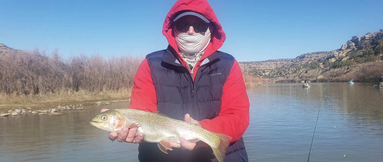 A man in a red hooded sweatshirt, a blue puffer vest, sunglasses, and a face mask holds up a freshly caught rainbow trout by a river on a clear sunny day. In the background, other people are fishing from boats on the water, with a dry, rocky hillside and sparse vegetation on the opposite bank.