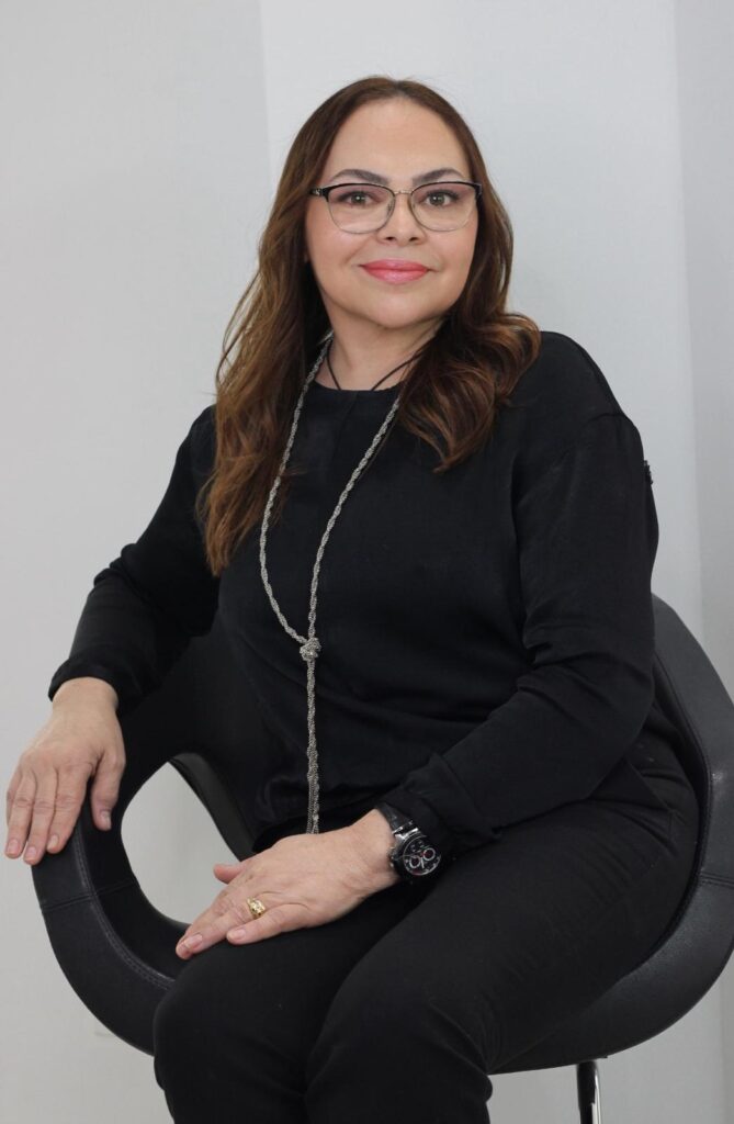 A woman with brown hair and glasses is seated in a chair, wearing a black long-sleeved shirt, black pants, and a long silver necklace. She is also wearing a watch on her left wrist and a ring on her right hand. The background is a plain white wall.