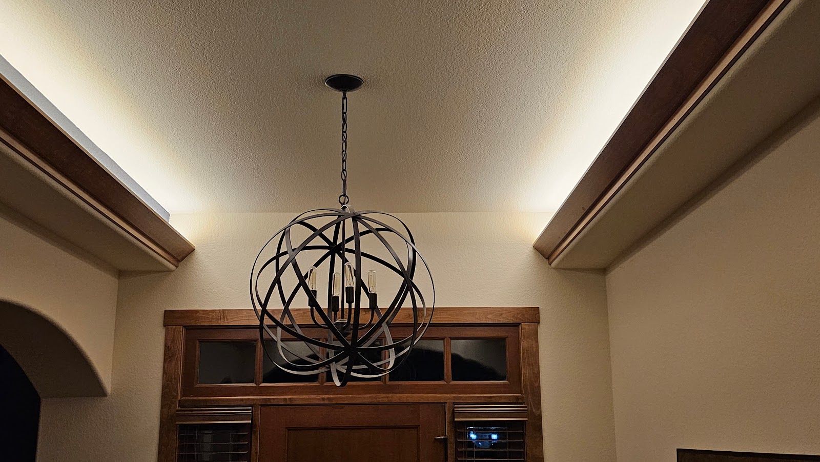 A black orb-shaped chandelier hangs from a chain against a textured ceiling. Recessed lighting illuminates the ceiling edges, which are accented with dark wood trim. Below the chandelier is a wooden door with glass panes and horizontal blinds.