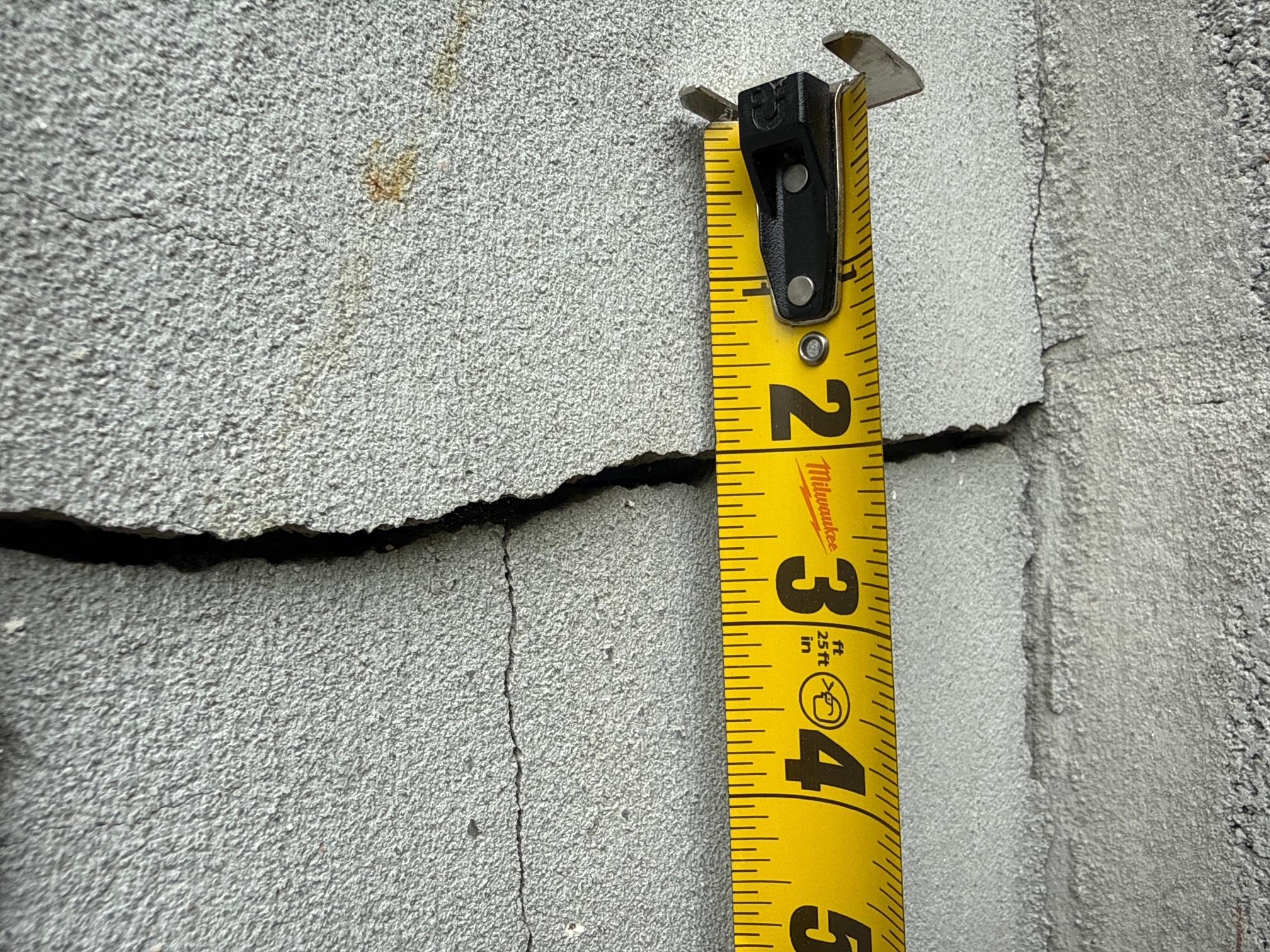 A Milwaukee tape measure hooked into a diagonal crack in a concrete masonry block, measuring its width.