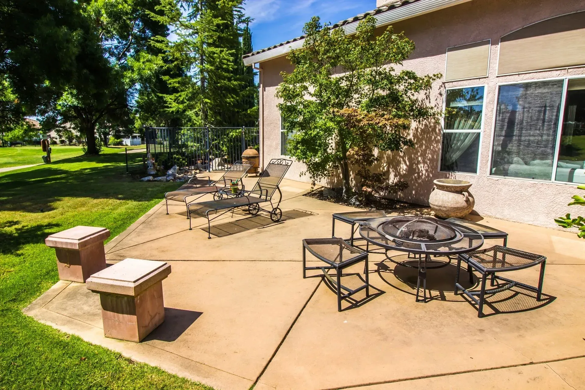 Well-maintained backyard patio featuring lounge chairs, fire pit table, landscaped greenery, and spacious concrete surface, creating an inviting outdoor extension for relaxation and entertainment.