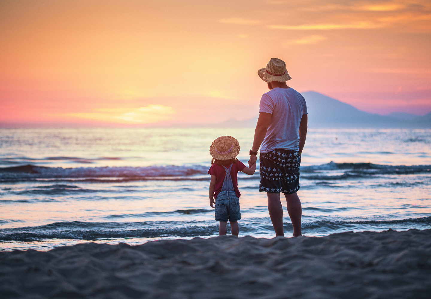 A father and young child holding hands on the beach and looking out at the water during sunset.