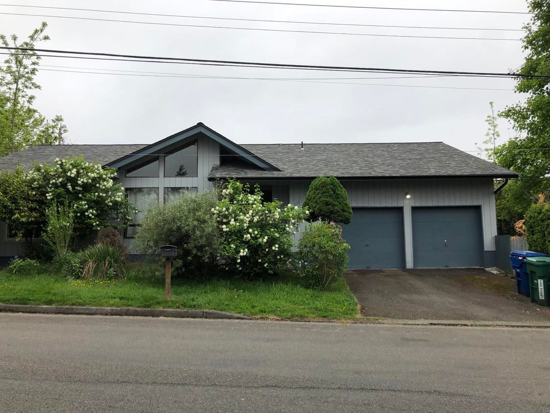 A gray ranch-style home features vertical siding, dark trim, a broad shingle roof, attached double g 