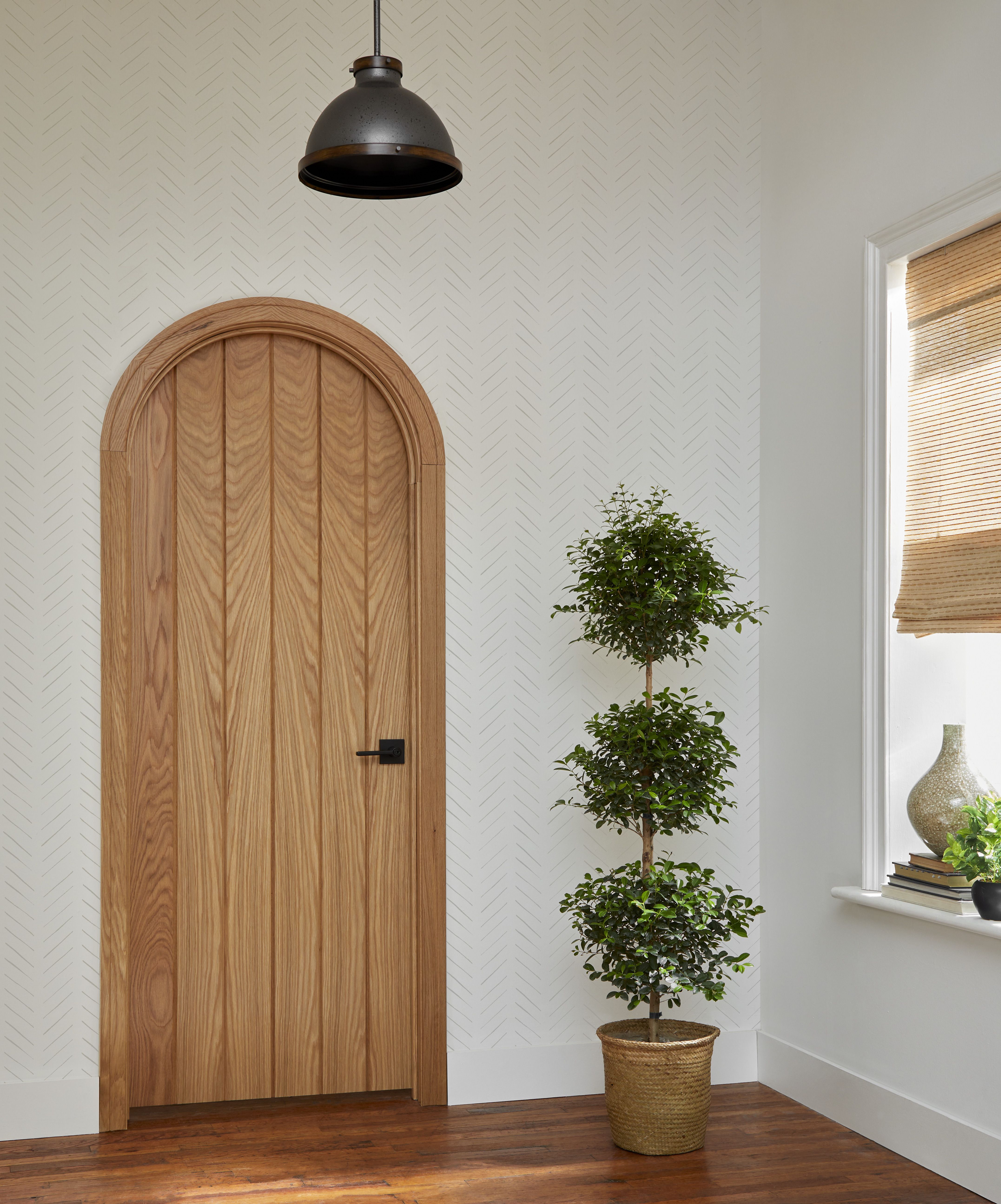 Custom arched interior wood door with natural grain detailing in a bright room featuring white patterned walls, hardwood floors, a hanging pendant light, and a decorative potted plant.
