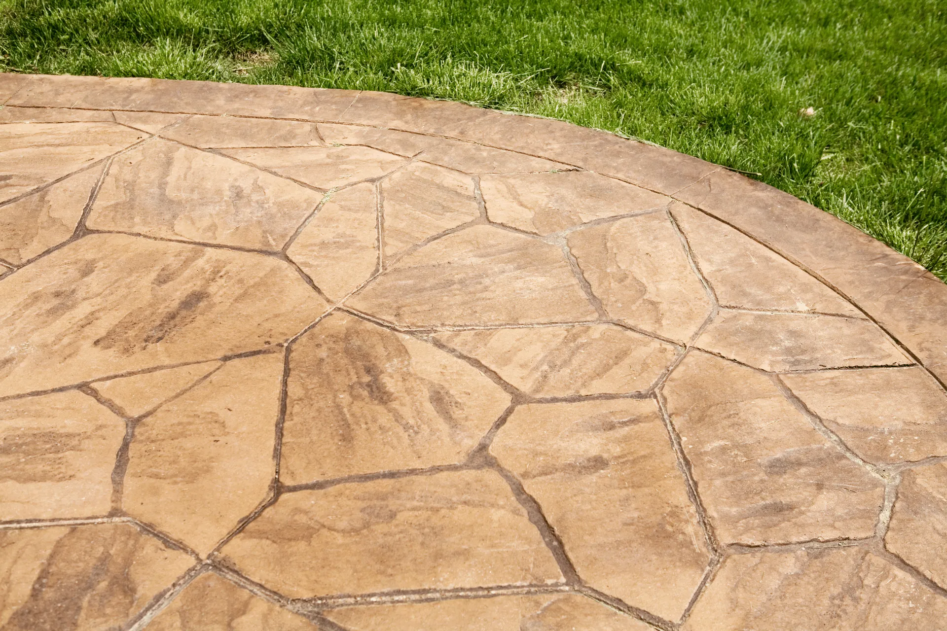 A close-up view of a stamped concrete patio with a flagstone pattern, bordered by lush green grass. The concrete is a warm tan color with darker lines defining the irregular shapes of the faux stones. The grass is vibrant and well-maintained, creating a natural contrast with the man-made patio.