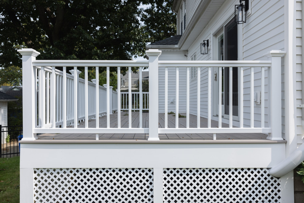 A white wooden deck railing with a lattice skirting is shown in front of a white vinyl-sided house. The deck floor is gray wood. Two black outdoor light fixtures are mounted on the house wall. A sliding glass door is visible in the background.