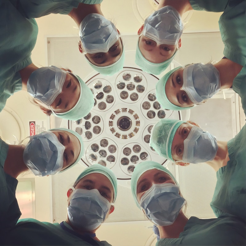 A group of medical professionals in scrubs, surgical caps, and masks are looking down at the camera from a low angle, gathered around a surgical light fixture. The image is taken from their perspective, looking upwards.