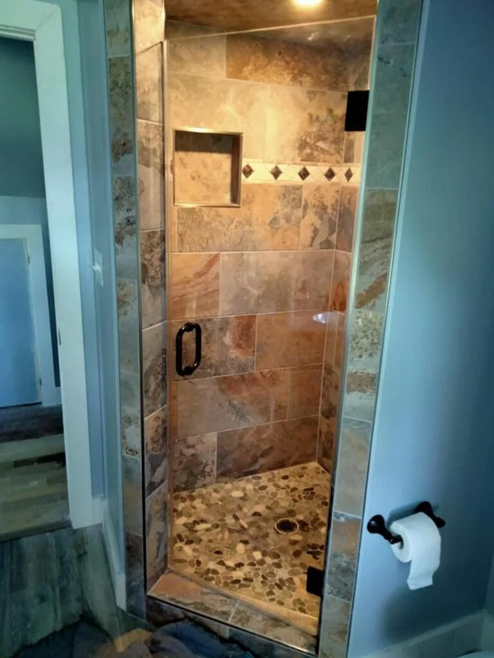A modern bathroom with a glass-enclosed shower featuring a stone tile floor and walls, as well as a recessed shelf. A roll of toilet paper is mounted on the light blue wall next to the shower.