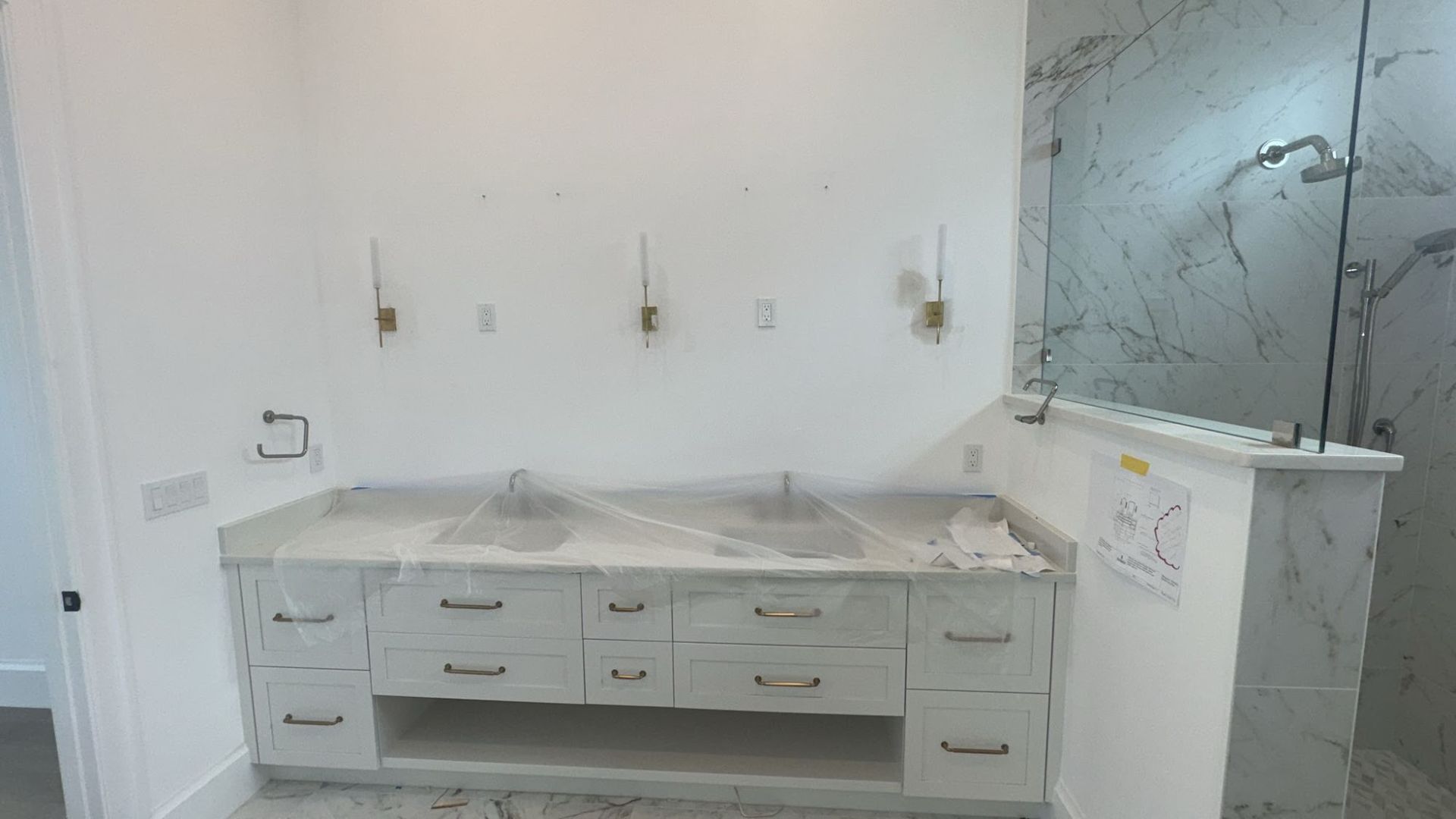 Newly installed double bathroom vanity with white cabinetry, marble countertop, and brass hardware, partially covered with protective plastic, positioned beside a glass shower enclosure with marble tile walls.