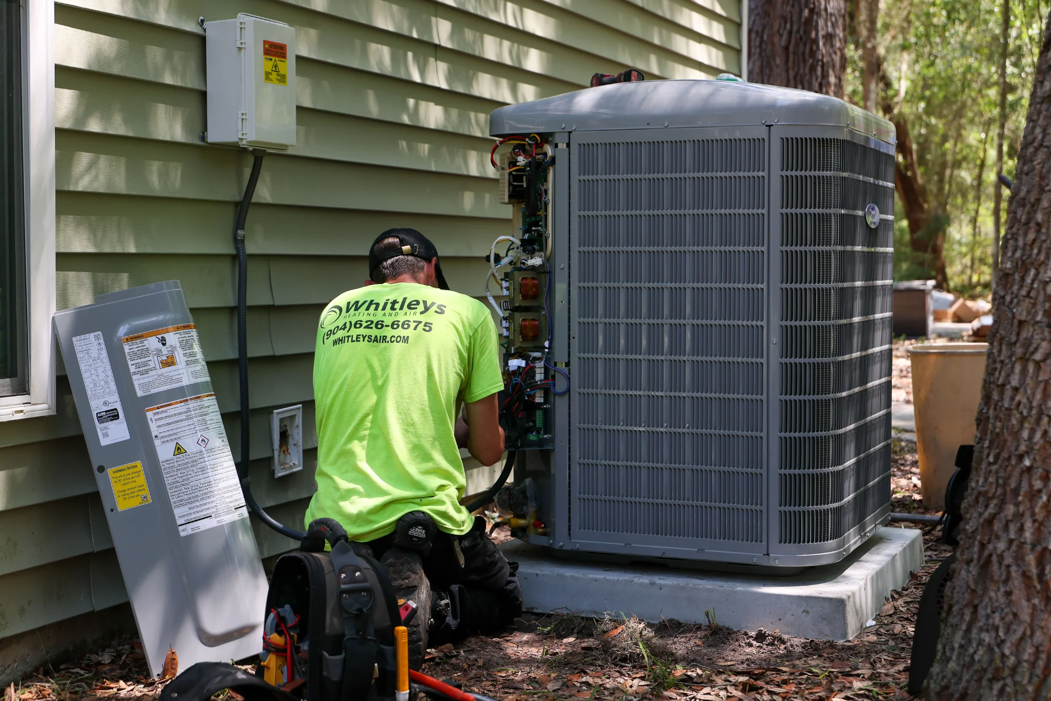 A technician in a bright green shirt with a black baseball cap kneels in front of an outdoor air con 