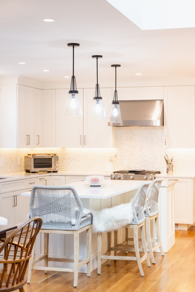 A bright and modern kitchen island with three pendant lights hanging above. The island has a white countertop and is adorned with two bar stools. One stool is covered with a fluffy white sheepskin throw. A glass bowl containing pink items sits on the countertop. In the background, there are white cabinets, a stainless steel range hood, and a toaster oven.