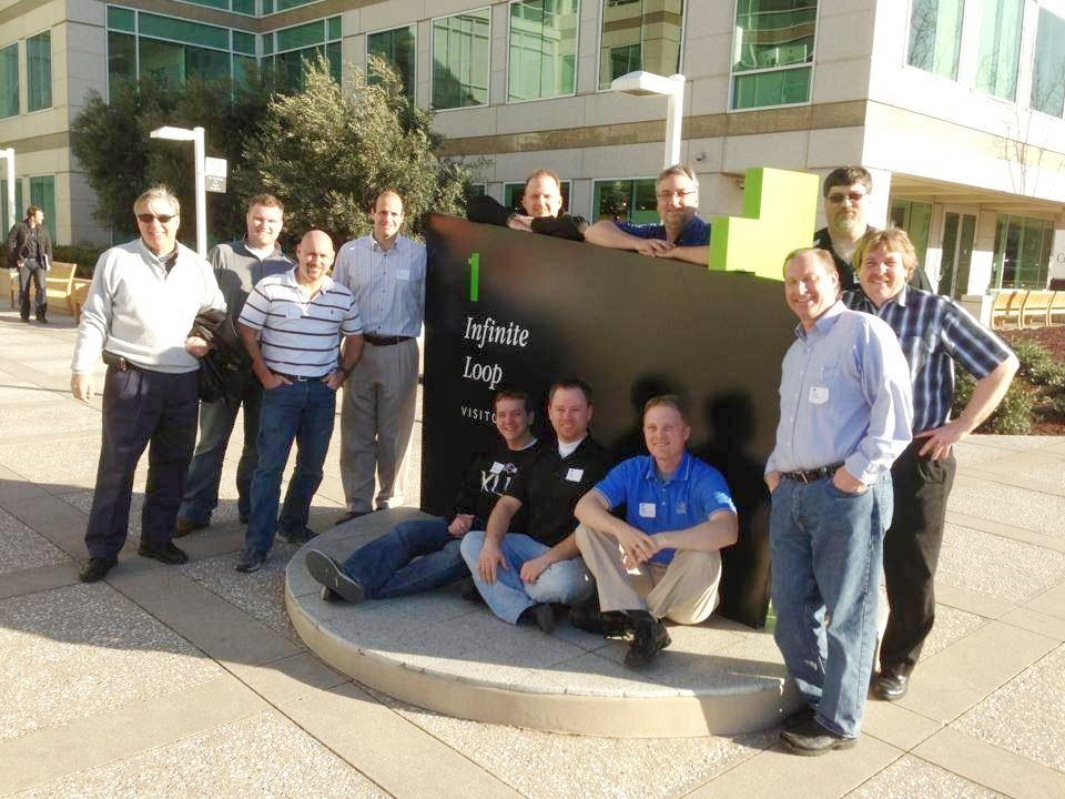 A group of ten men pose for a photo in front of a large black sign that reads "1 Infinite Loop Visitor". The sign is located in front of a modern, multi-story building with a glass facade. Several of the men are standing, while two are sitting on a raised circular platform in front of the sign. They are all dressed casually in shirts and pants, and some are wearing lanyards with identification badges. The sun is shining, casting shadows on the ground, suggesting it is daytime.