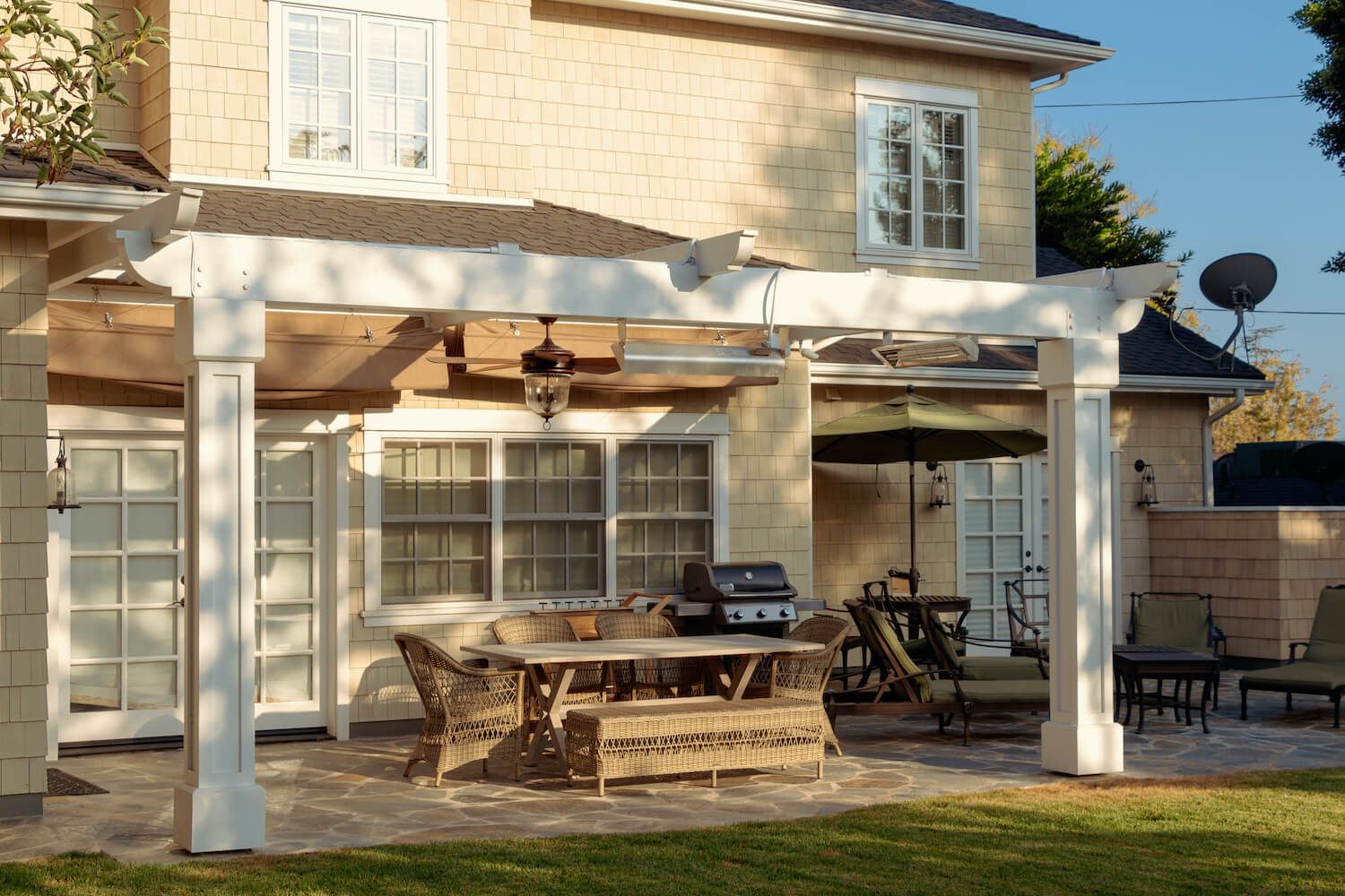 Sunlit backyard patio with wicker dining set, grill, and lounge chairs beneath a covered terrace beside a bright, welcoming home.