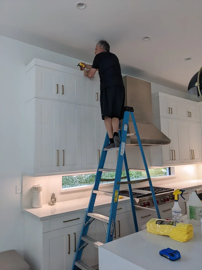 A man in a black t-shirt and shorts stands on a blue ladder in a modern kitchen, reaching up to adjust something near the ceiling. He holds a yellow tool in his right hand. The kitchen features white cabinets, stainless steel appliances including a range hood, and white countertops. A narrow window above the countertop offers a view of greenery. Several cleaning supplies and yellow cloths are visible on the countertop.