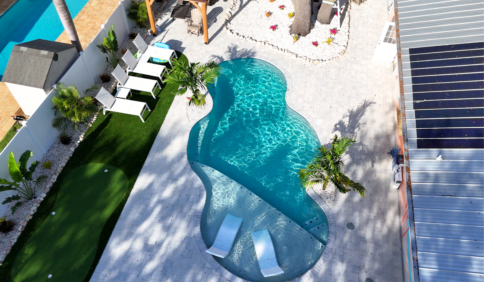 Aerial view of a custom freeform backyard swimming pool with an attached spa, spacious patio, lounge seating, and landscaped surroundings.