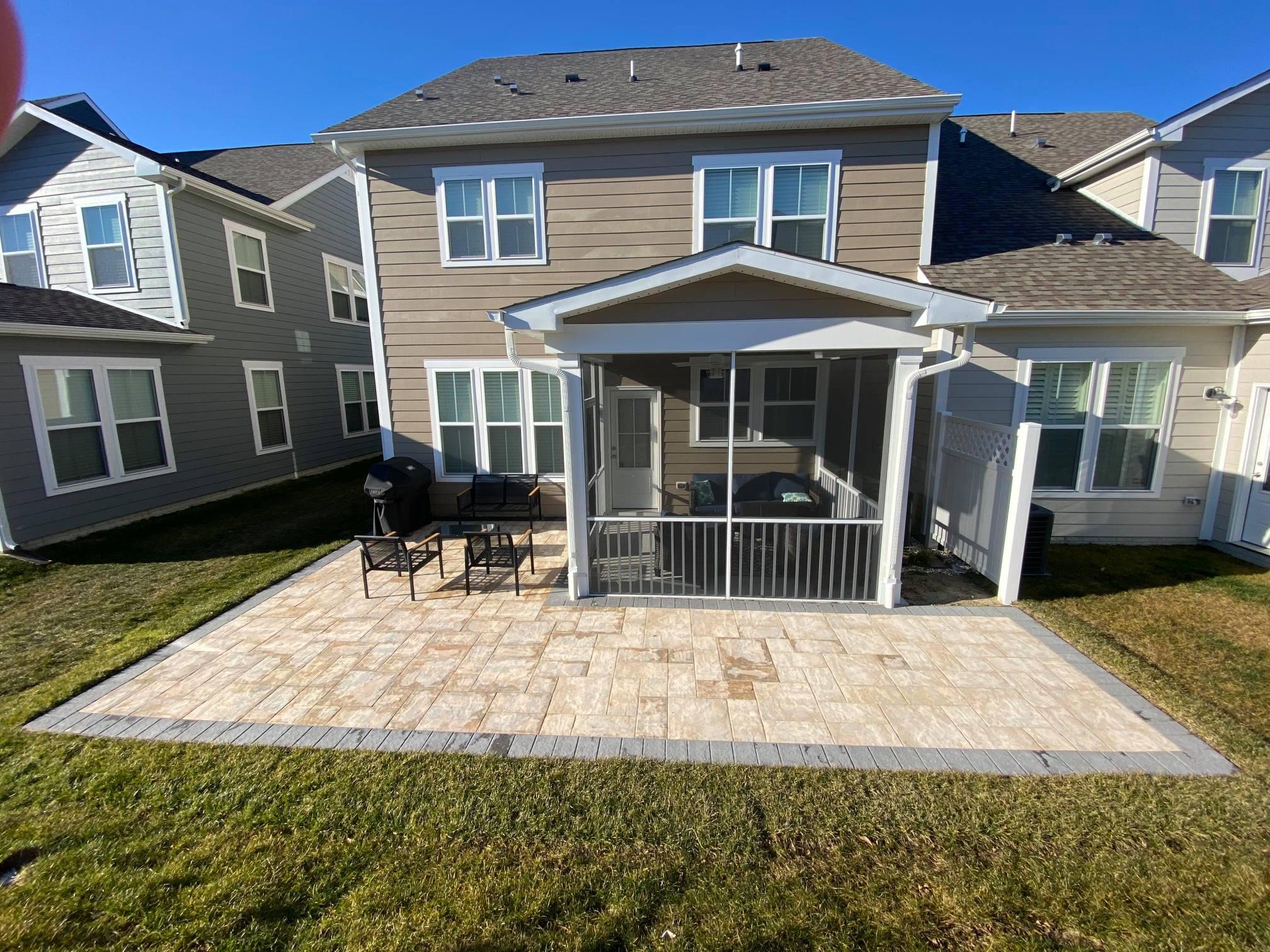 A professional hardscaping project underway in a Delaware home's backyard. The image shows landscapers laying irregular stone pavers to create a winding pathway. The scene includes a variety of landscaping tools and materials like sand and stone, with a partially completed patio area visible in the background. The house and garden display early spring foliage.