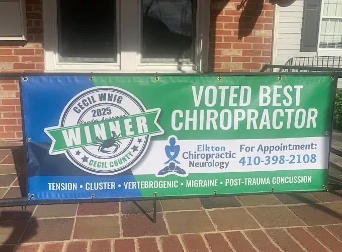 It&rsquo;s a photo of a large outdoor promotional banner mounted along a porch/railing in front of a brick building.
The banner has a blue-to-green gradient background. On the left is a circular badge that reads &ldquo;CECIL WHIG 2025 Reader Approved WINNER &ndash; Cecil County.&rdquo; On the right, large text says &ldquo;VOTED BEST CHIROPRACTOR.&rdquo;
In the middle/right area it shows the Elkton Chiropractic Neurology name and logo (a meditating figure icon), and it lists &ldquo;For Appointment: 410-398-2108.&rdquo; Along the bottom, it includes conditions/topics such as tension, cluster, vertebrogenic, migraine, and post-trauma concussion.