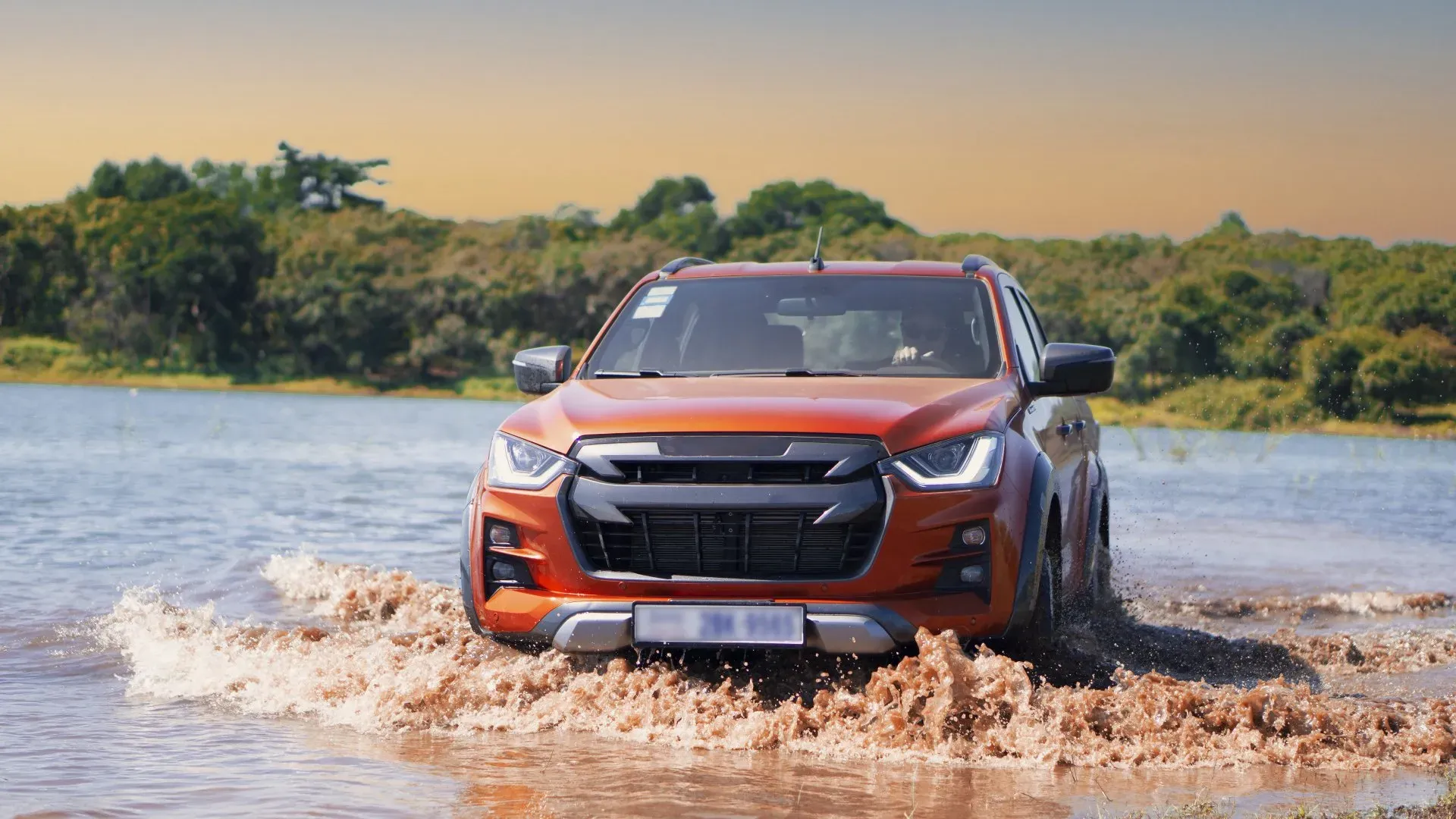 A bright orange Isuzu D-Max pickup truck drives through shallow, muddy water. The truck is captured from a low angle, emphasizing its front grille and sharp LED headlights. Splashes of mud and water erupt around the vehicle, highlighting its off-road capability. In the background, a body of water stretches towards a lush green treeline under a soft, golden-orange sky.