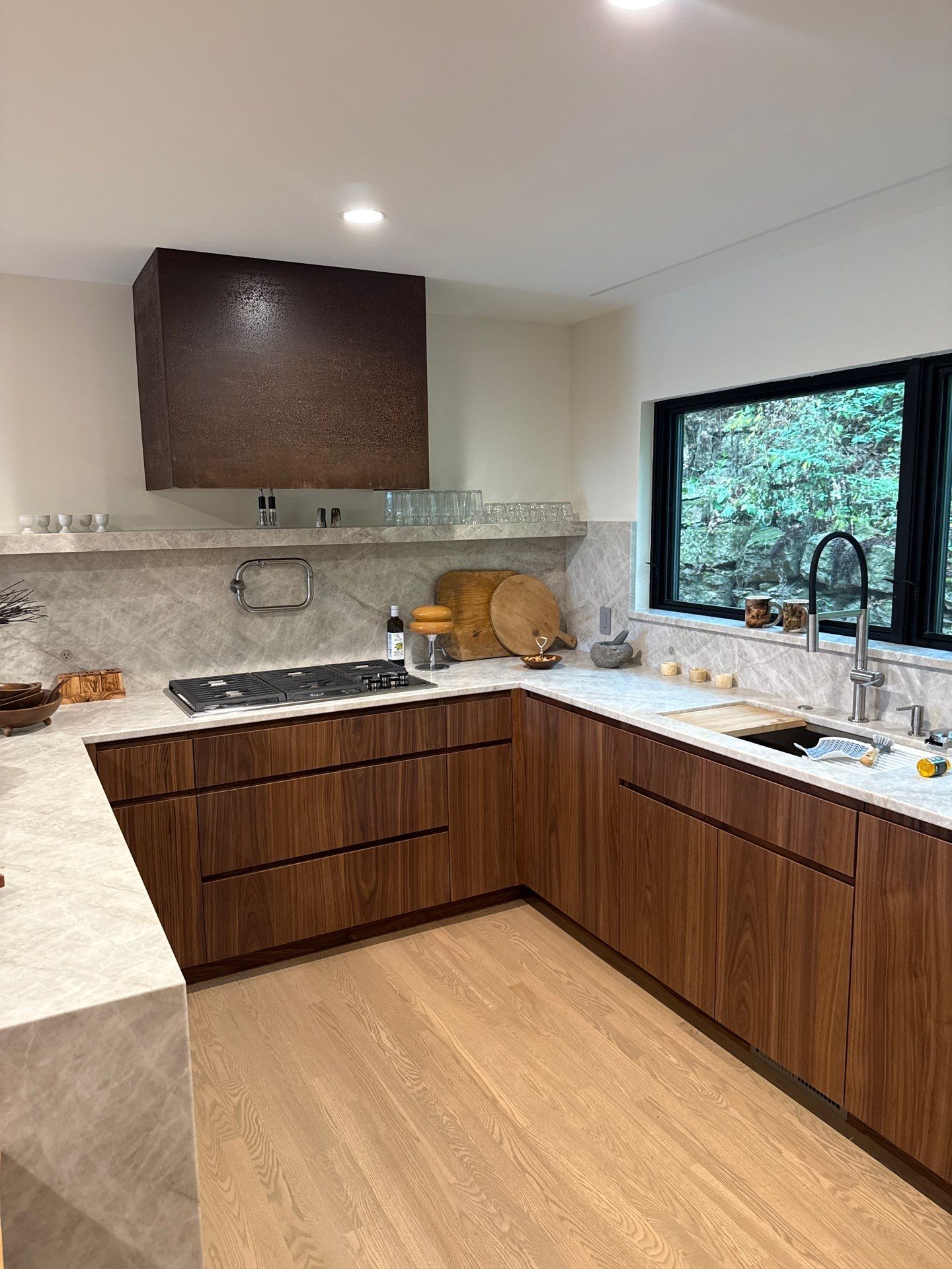 Modern kitchen features flat-panel walnut cabinetry, pale stone countertops and backsplash, black sink with tall faucet, gas cooktop, large dark range hood, wide window, and light wood flooring throughout indoors.