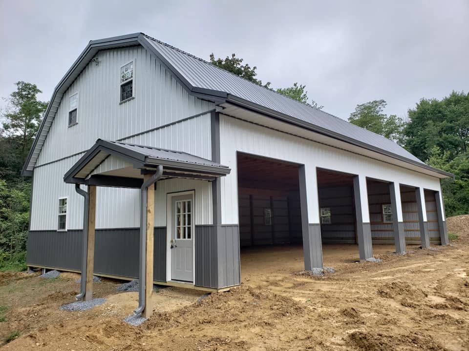 A newly constructed metal barn-style building with white siding and gray trim, featuring multiple open bays, a covered entrance, and a sloped roof, set on a dirt lot surrounded by trees.