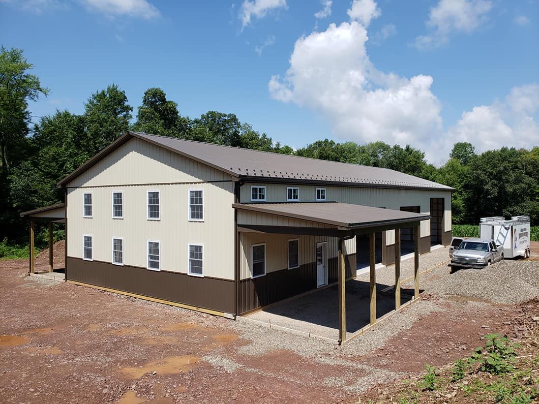 A large metal building with beige siding and brown trim, featuring multiple windows, a covered porch area, and attached garage bays, set on a gravel lot surrounded by trees.