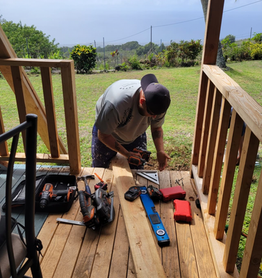 A construction worker, wearing a cap and gray shirt, is cutting a piece of wood with a rotary saw on a deck. Various power tools and a level are laid out nearby on the deck planks.