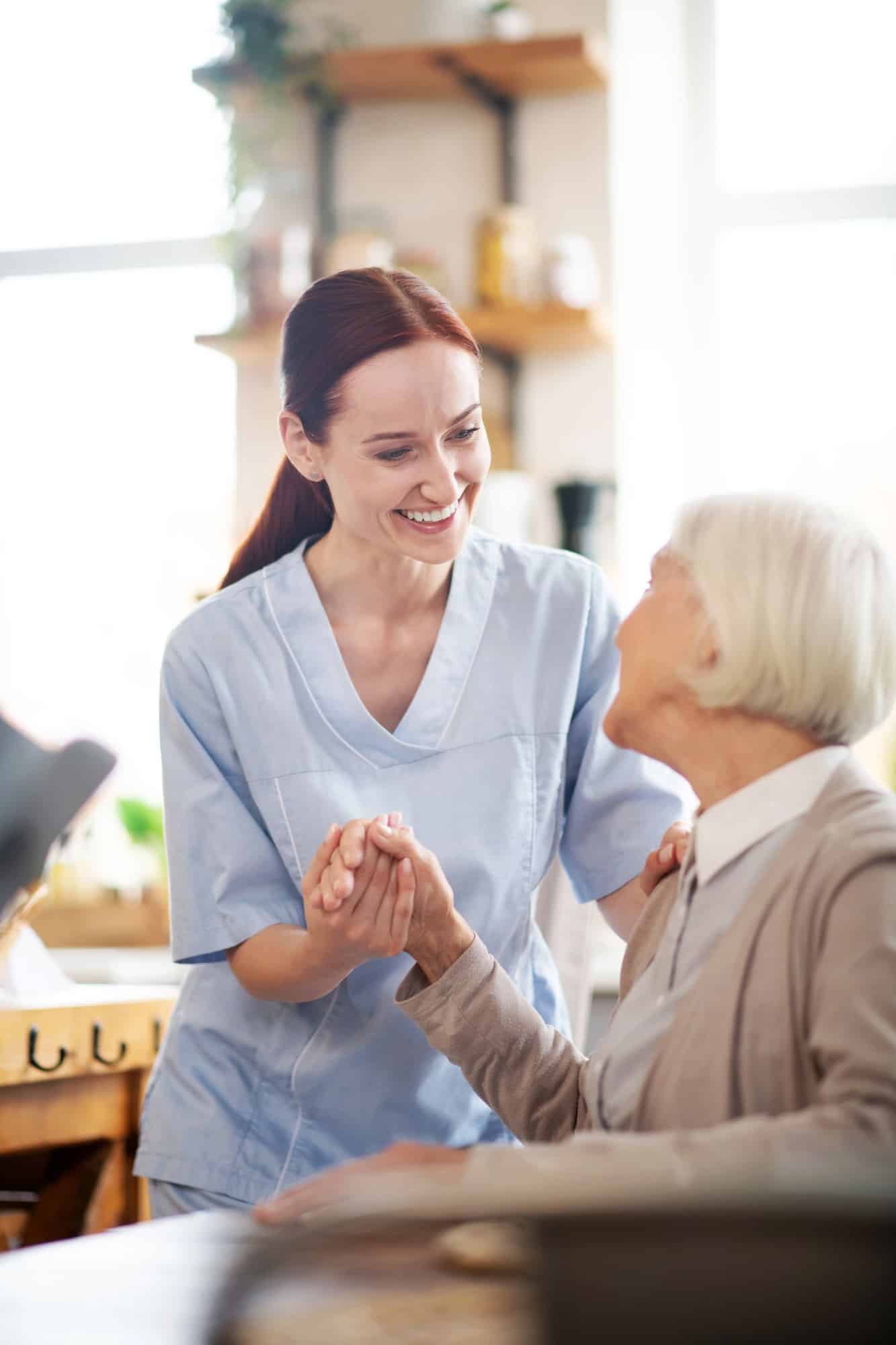 A caregiver in a light blue uniform holds the hands of an elderly woman and they both smile at each other. The caregiver is leaning slightly forward, and the elderly woman is sitting, looking up at the caregiver. The background is blurred, but appears to be an indoor setting, possibly a home or care facility, with shelves and a window visible.