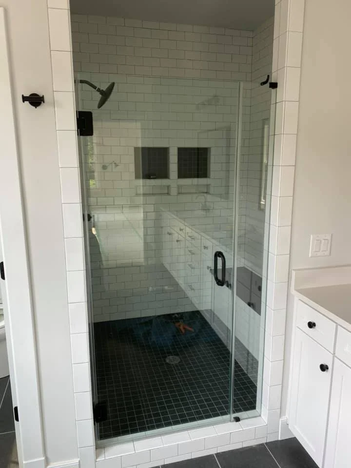 A modern bathroom features a glass-enclosed shower with white subway tile walls and a dark tile floor. A chrome showerhead is visible inside the shower. To the right, a white vanity with black knobs is partially visible, along with a white countertop.