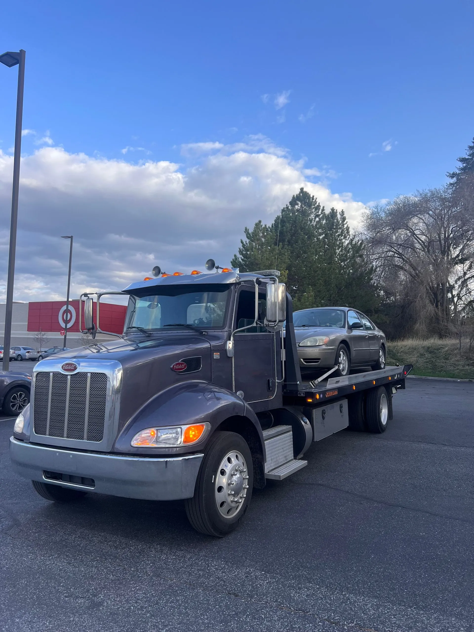 Direct Towing flatbed truck transporting a sedan near a commercial retail area, highlighting professional towing services, secure vehicle transportation, roadside assistance expertise, and dependable recovery operations.