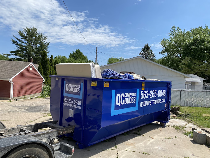 A blue QC Dumpster Dudes dumpster filled with furniture and miscellaneous items, located in a residential driveway.