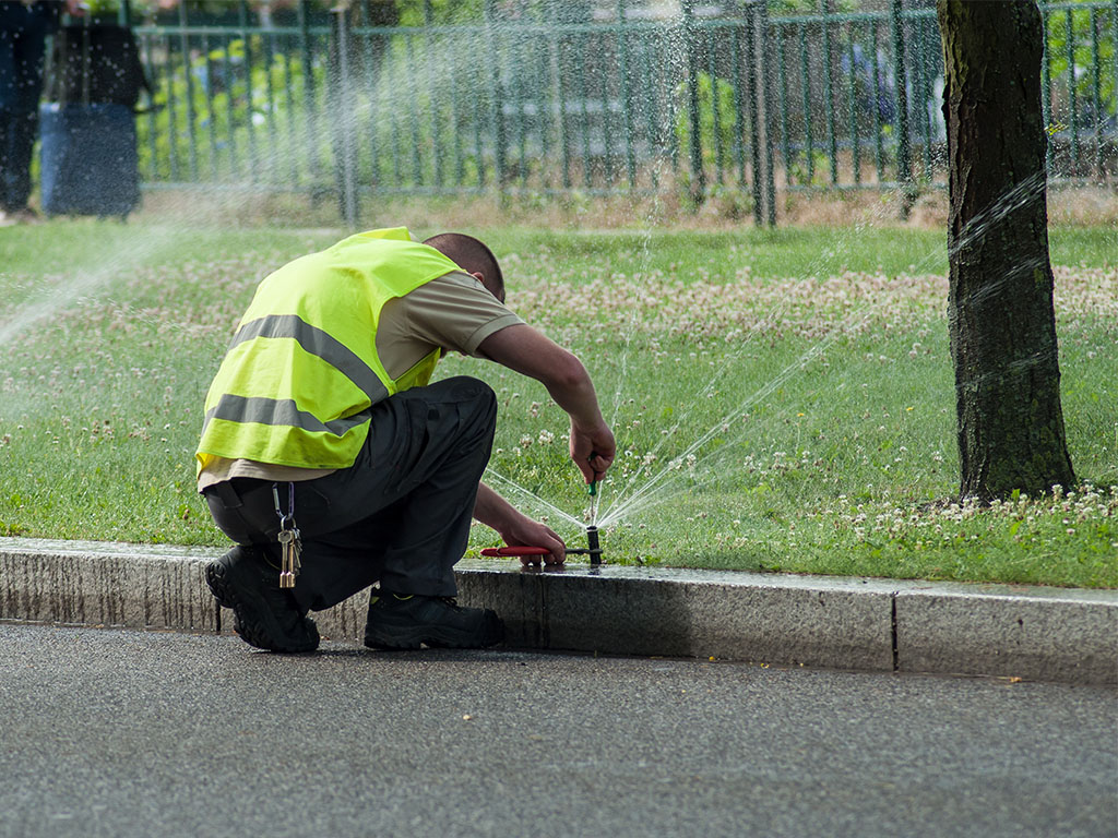 A technician in a high-visibility safety vest kneeling to adjust an active lawn sprinkler head next to a curb.