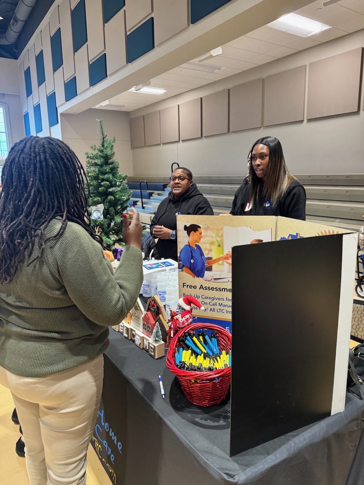 A woman with dreadlocks stands at a table speaking with two women behind it. A small decorated Christmas tree is on the table, along with bottles of hand sanitizer, decorative houses, a basket of pens, and a sign. The woman speaking to the two women wears a green sweater and beige pants. The two women behind the table are wearing black hoodies with logos. The woman on the left wears glasses. The woman on the right has long dark hair with blonde highlights. A large sign with an image of a nurse and text is visible on the table.