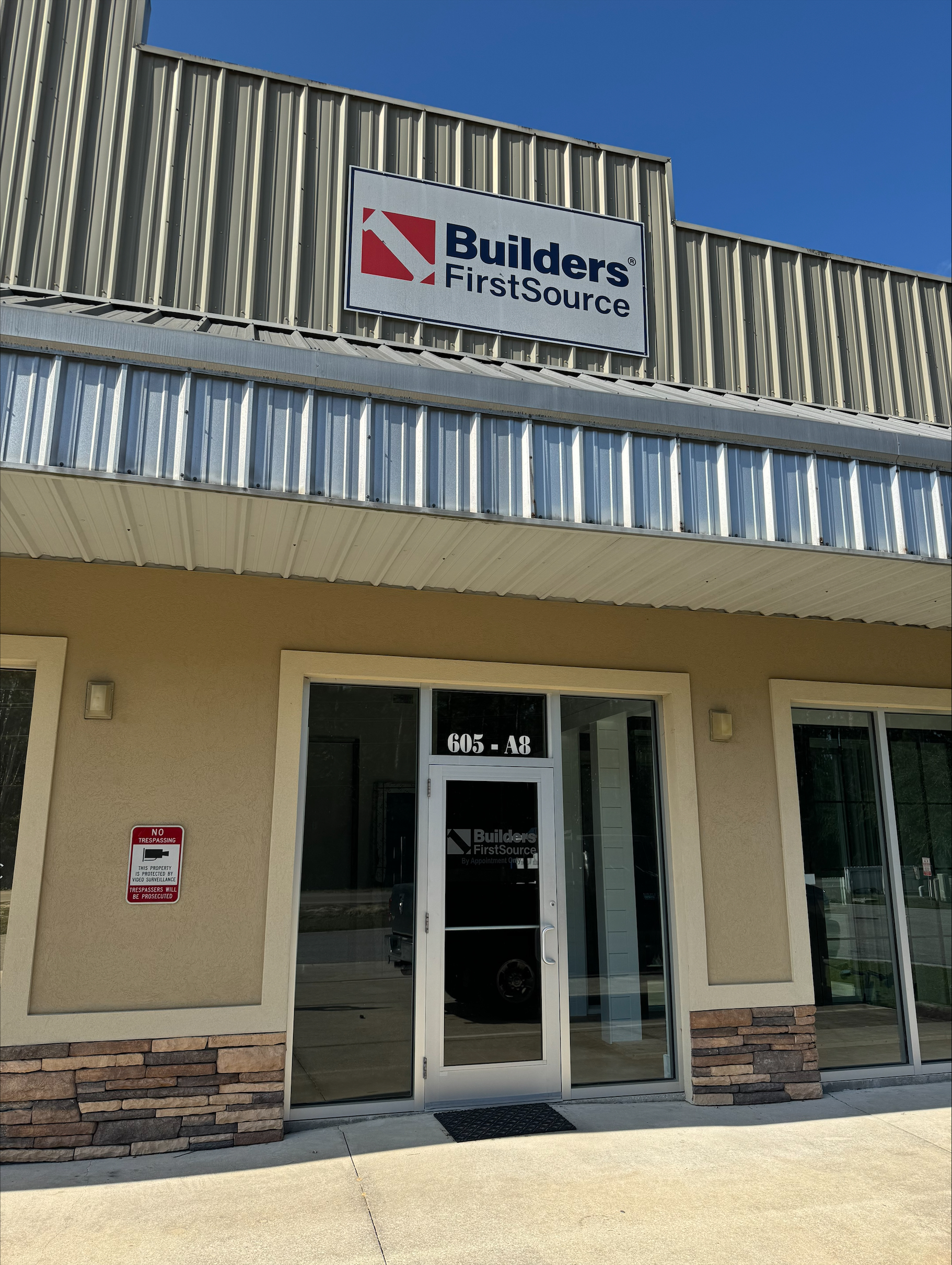 A Builders FirstSource storefront in a beige building with stone accents, featuring a prominent company sign above the entrance. The glass door displays the company logo, and the address "605 - A8" is visible above it. A bright blue sky adds to the clean and inviting appearance of the location.