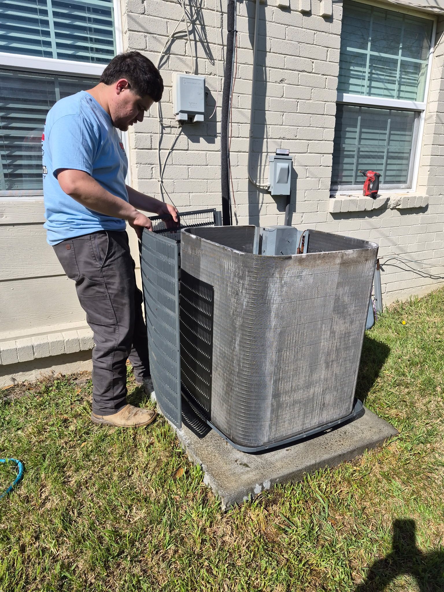 Technician removing panel from outdoor condenser unit for inspection or repair. Residential HVAC maintenance in progress, showing cleaning, troubleshooting, or component replacement beside house exterior wall.