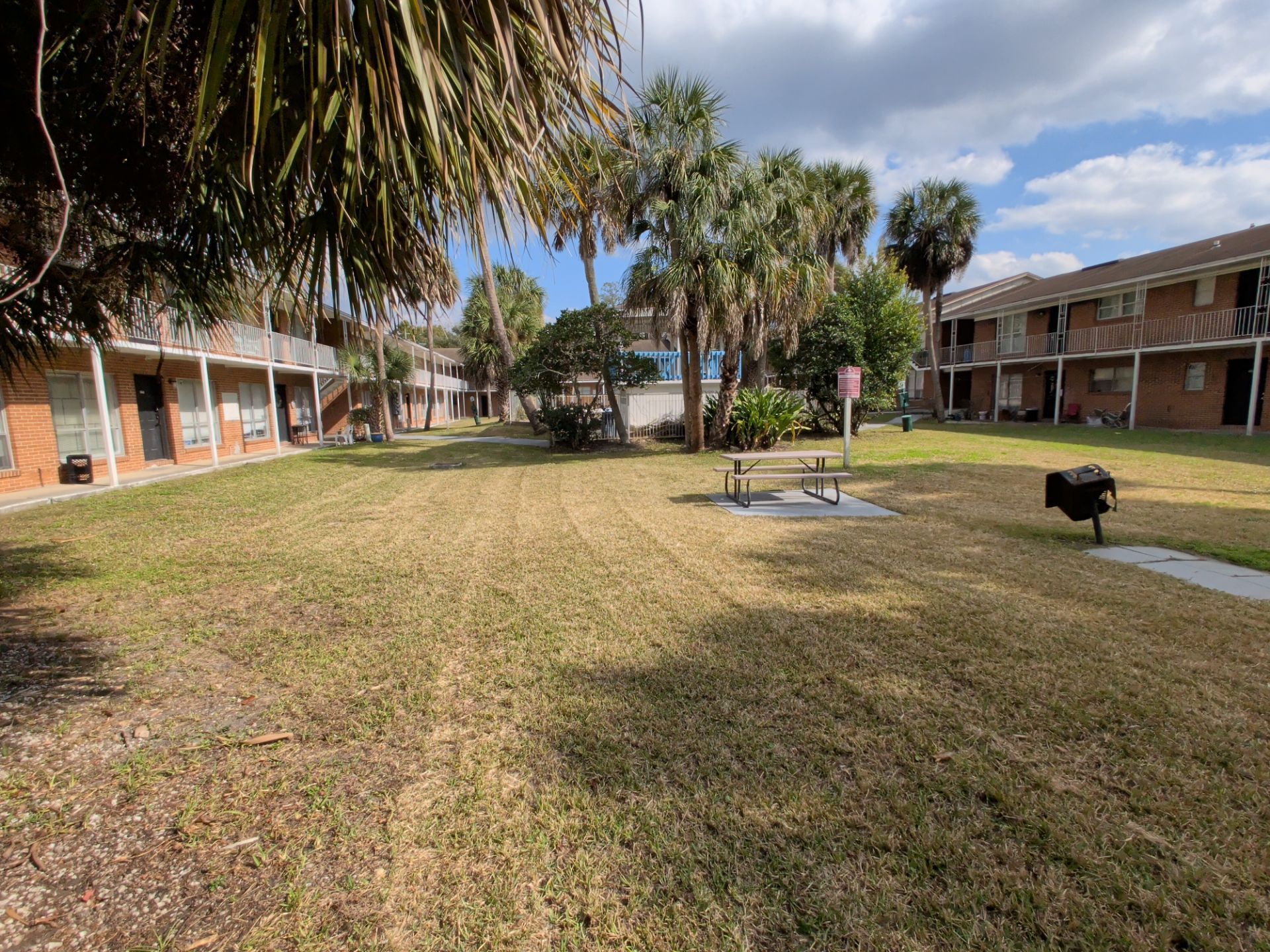 Apartment complex courtyard with grassy lawn, picnic table, and barbecue grill. Palm trees and landscaped plants surround shared outdoor space between two-story brick residential buildings with balconies.