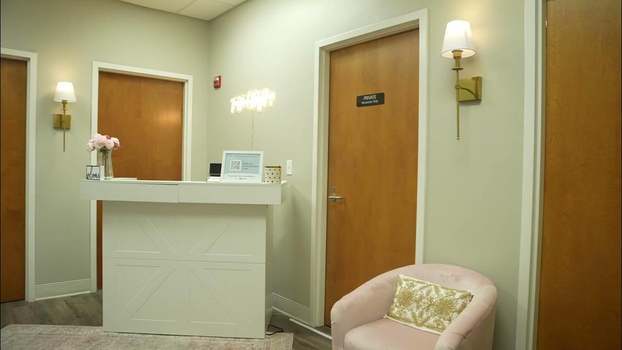 A brightly lit, modern reception area with a white counter, a pink armchair, and two wooden doors. The walls are painted a light green. On the counter, there's a vase of pink flowers, a framed QR code, and a laptop. A sign on one of the doors reads "PRIVATE".