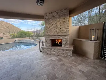 Scottsdale patio remodel with a custom outdoor wood-burning fireplace, stacked stone surround, and travertine flooring throughout. Pool visible in the background, Pinnacle Peak beyond the wall. The covered patio ties the fireplace and pool deck into one continuous outdoor living space. Built by Hacienda Outdoors, ROC #325952.
