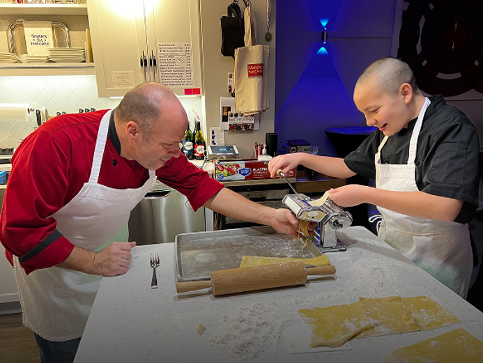 An adult man in a red chef's jacket and a white apron works with a young person in a black chef's jacket and a white apron, both making fresh pasta using a pasta machine on a floured countertop. The man is guiding the dough, while the young person operates the crank. Several sheets of pasta dough, a rolling pin, and a fork are visible on the counter, along with a metal baking pan. The kitchen setting includes shelves with dishes and bottles, and a modern blue light fixture on the wall.