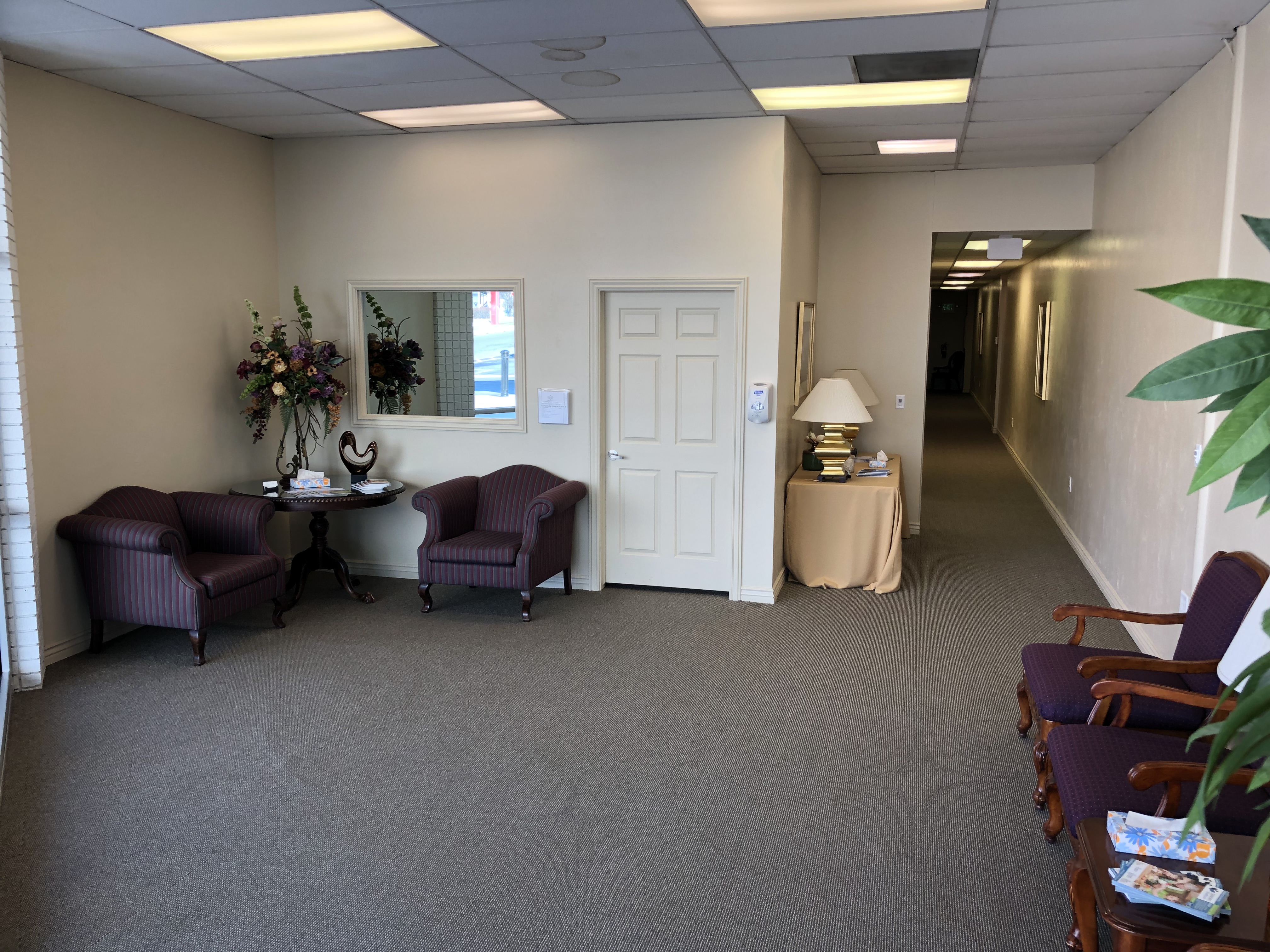 Reception area opening onto a carpeted corridor, with armchairs, a mirrored console holding fresh flowers and a lamp-lit table outside the offices.