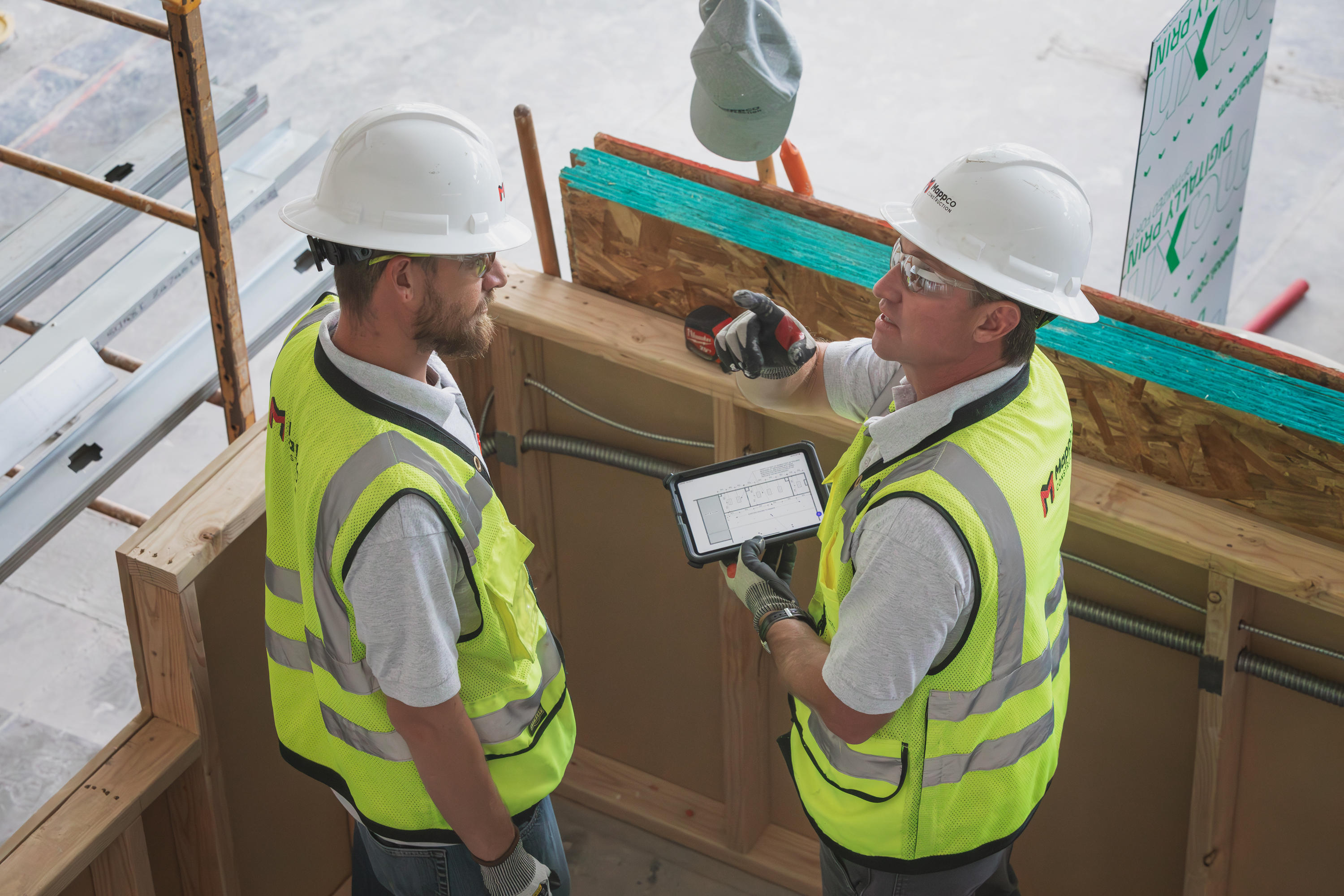 Two Mappco Construction professionals are in the midst of a project site consultation. Chad Mapp, on the right, is holding a tablet, displaying blueprints or project plans, while gesturing towards an area of interest. They stand within a partially completed structure, surrounded by building materials and insulation layers.