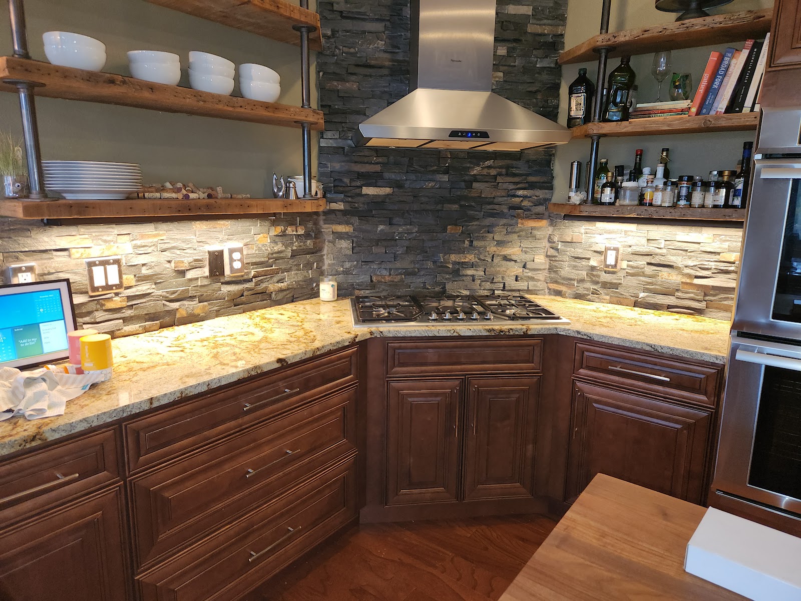A kitchen counter with a gas stovetop, granite countertops, and a stone tile backsplash. Above the counter are wooden shelves displaying bowls, plates, wine bottles, and books. A stainless steel range hood is mounted above the stovetop. To the right, part of a stainless steel oven is visible.