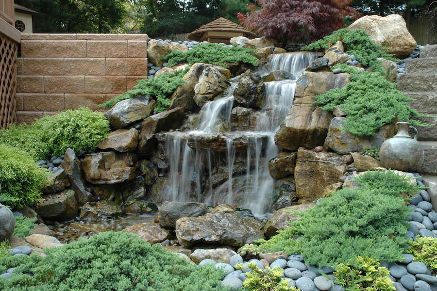 Lush Japanese garden waterfall cascades over layered rocks into a stream, surrounded by mossy shrubs, smooth stones, and a pottery jar.