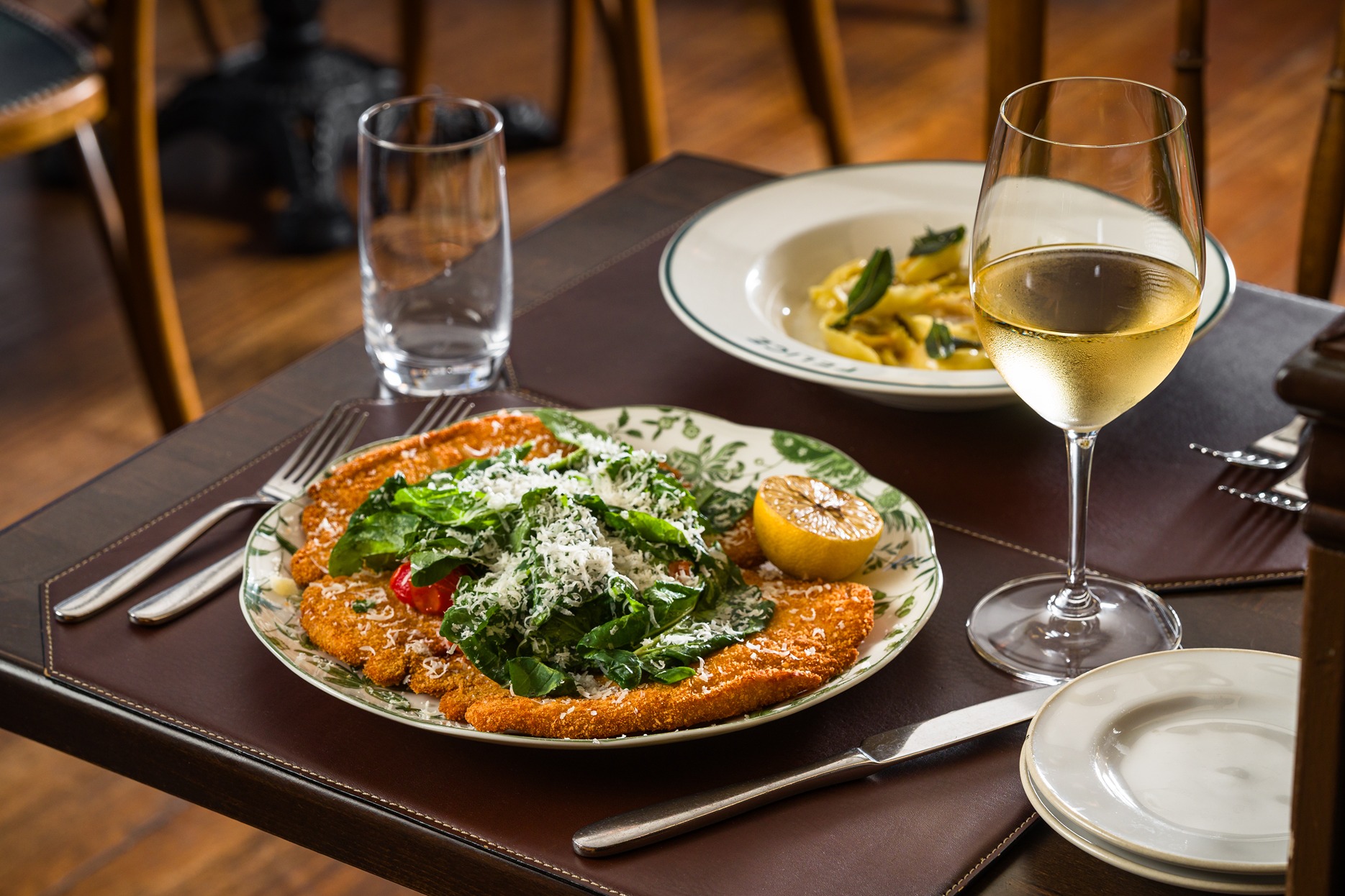 A crispy veal milanese topped with a salad of greens and shaved cheese, served on a floral plate with a grilled lemon and a glass of white wine on a leather tabletop.